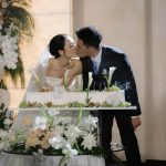 Cô dâu chú rể bên bàn bánh cưới trang trí hoa và nến.